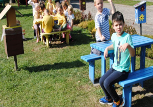 dzieci siedzą i stoją obok ławek i stolików w ogrodzie wg ich kolorów i ubrań (żółty i niebieski))